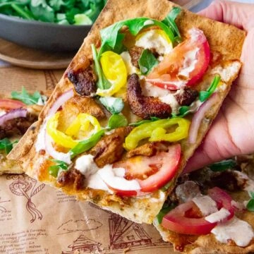 Shawarma pizza topped with tomatoes, yellow peppers, onions, and greens, held by hand over decorative paper.