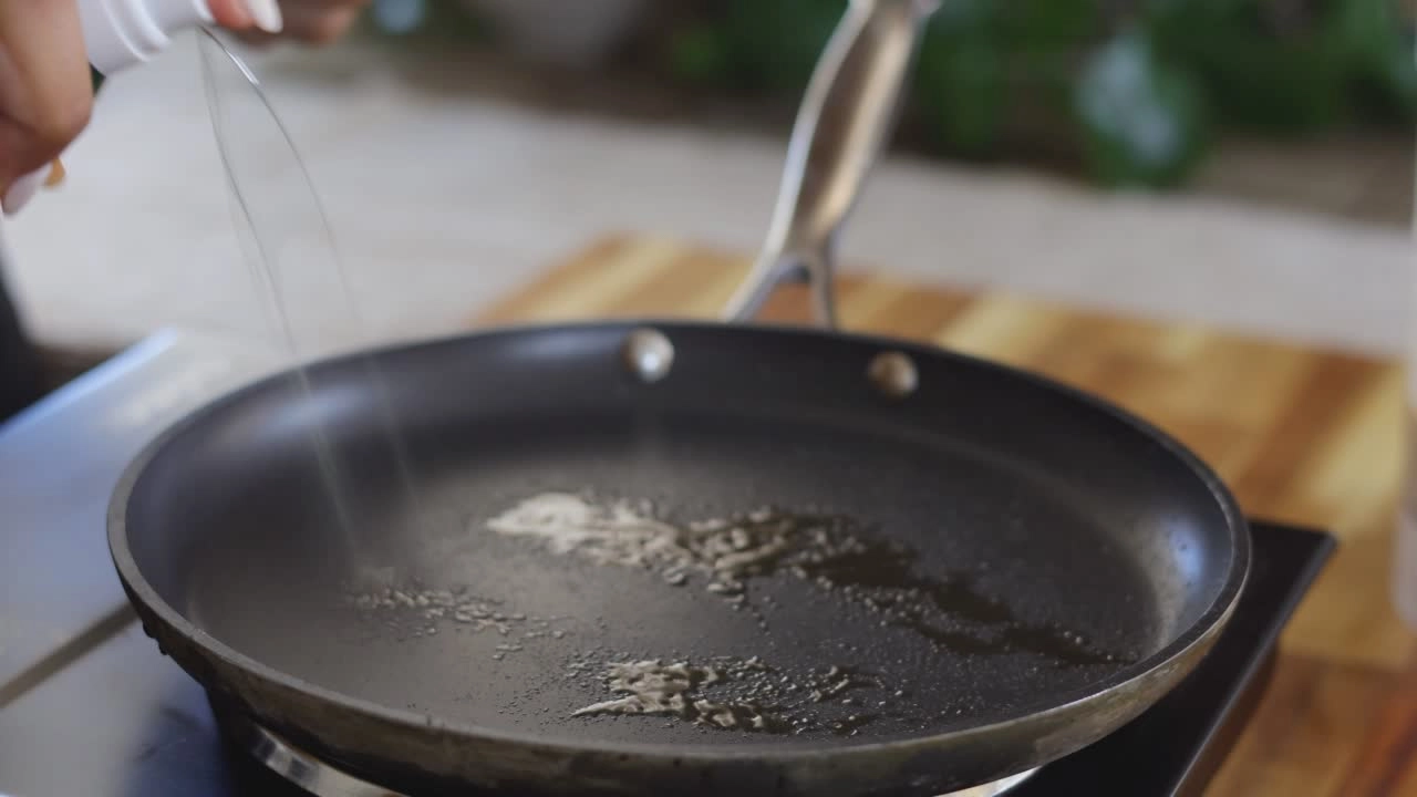 Pouring oil into a black frying pan on a kitchen stove for cooking preparation.