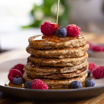 Stack of banana oatmeal pancakes topped with syrup, raspberries, and blueberries on a plate.