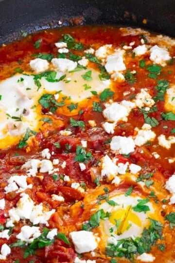 Skillet of shakshuka with poached eggs, tomatoes, feta cheese, and herbs. Savory Mediterranean breakfast dish.