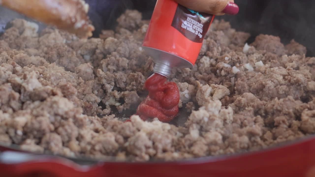 Adding tomato paste to sizzling ground meat in a pan.