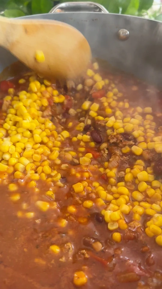 Cooking a chili in a pot with corn, beans, and tomatoes being stirred with a wooden spoon.