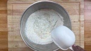 Bowl of flour with water being poured from a white measuring cup on a wooden surface. Perfect for baking or cooking.