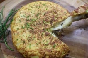 Cheesy Spanish tortilla with diced chives on a wooden board, showcasing a delicious slice being lifted.