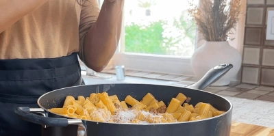 Person cooking rigatoni pasta with cheese in a pan by a sunny kitchen window.