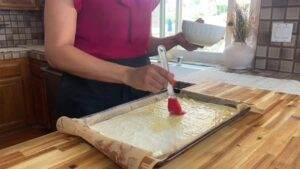 Person brushing olive oil on filo pastry sheet in a kitchen. Baking preparation on wooden countertop with natural light.