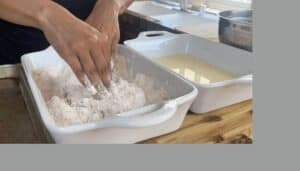 Hands mixing flour in a white baking dish, preparing dough on a wooden kitchen counter.
