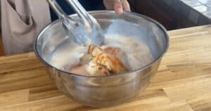 Marinating chicken in a stainless steel bowl with buttermilk, held by kitchen tongs on a wooden countertop.