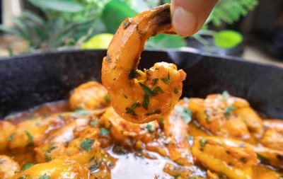 Close-up of hand holding a spicy shrimp topped with fresh herbs, cooked in flavorful sauce in a skillet.