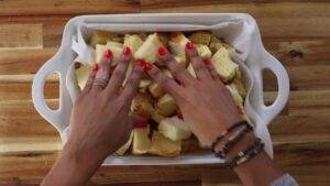 Hands arranging bread pieces in a white baking dish on a wooden table.