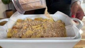 Sprinkling crunch topping on banana dessert in a white baking dish on a wooden table.