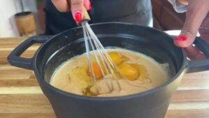 Whisking eggs into batter in a black pot on a wooden countertop, cooking preparation.
