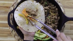 Person preparing beef fajita with bell peppers and avocado in a skillet.