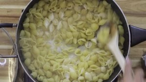 Cooking shell pasta in a pot with heavy cream, stirred with a wooden spoon on a wooden countertop.