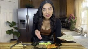Woman cooking in kitchen, holding a tortilla beside a skillet with vegetables and meat.