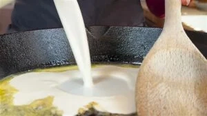 Pouring cream into a frying pan with a wooden spoon, cooking preparation close-up.