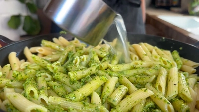 Pouring pasta water into pesto pasta in a skillet, creating a delicious pesto pasta.