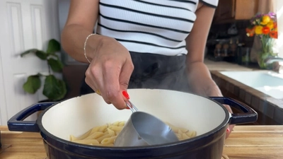 Person reserving pasta water from a white pot on a stove, wearing a striped shirt in a kitchen setting.