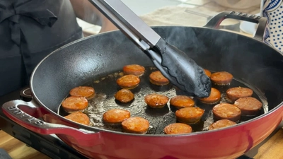 Chef cooking sliced sausage in a red pan with tongs, capturing the cooking process.