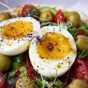 Mediterranean Hummus Bowl with Soft-boiled eggs on a vibrant salad with tomatoes, olives, and microgreens, sprinkled with spices.