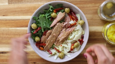Fresh salad with tuna, arugula, olives, sun-dried tomatoes, and fennel in a bowl on a wooden table.