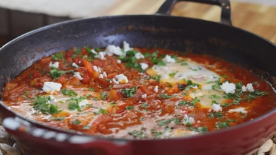 Pan of shakshuka with poached eggs in spicy tomato sauce, garnished with herbs and crumbled feta cheese.