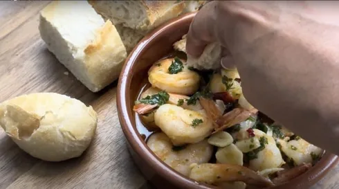 Hand dipping bread into a bowl of garlic shrimp with fresh herbs; served with crusty bread on a wooden table.