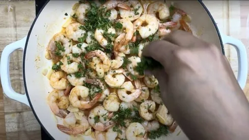 Person garnishing cooked shrimp with herbs in a white pan. Perfect for seafood recipe inspiration.