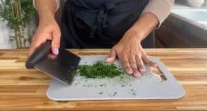 Person chopping fresh herbs on a cutting board in a kitchen setting.