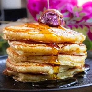 Stack of fluffy pancakes drizzled with syrup, topped with a berry, and set against a floral background.