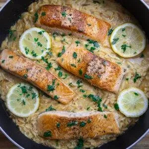 Pan-seared salmon with creamy lemon orzo, garnished with parsley and lemon slices in a skillet.