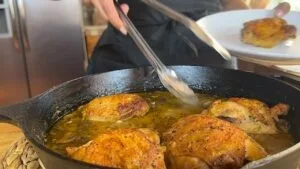 Chef adding crispy chicken to a skillet with tongs, rich sauce simmering below. Perfectly cooked, golden brown skin.