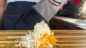 Person grating cheddar and mozzarella cheese on a wooden cutting board in a kitchen.