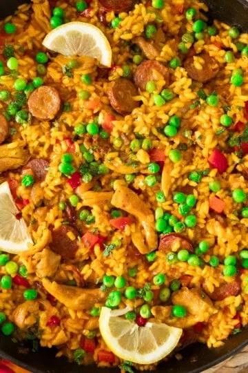 Pan of vibrant paella with saffron rice, peas, chorizo, chicken, red peppers, and lemon wedges.