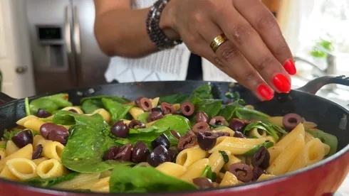 Fresh pasta dish with spinach and olives being garnished by a hand in a kitchen setting.