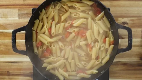 Steaming penne pasta with cherry tomatoes cooking in a black pot on a wooden surface.