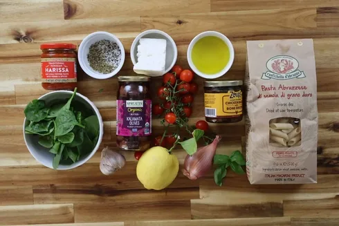 Ingredients for creamy veggie pasta. They are penne pasta, cherry tomatoes, olives, harissa, spinach, lemon, feta, and olive oil.